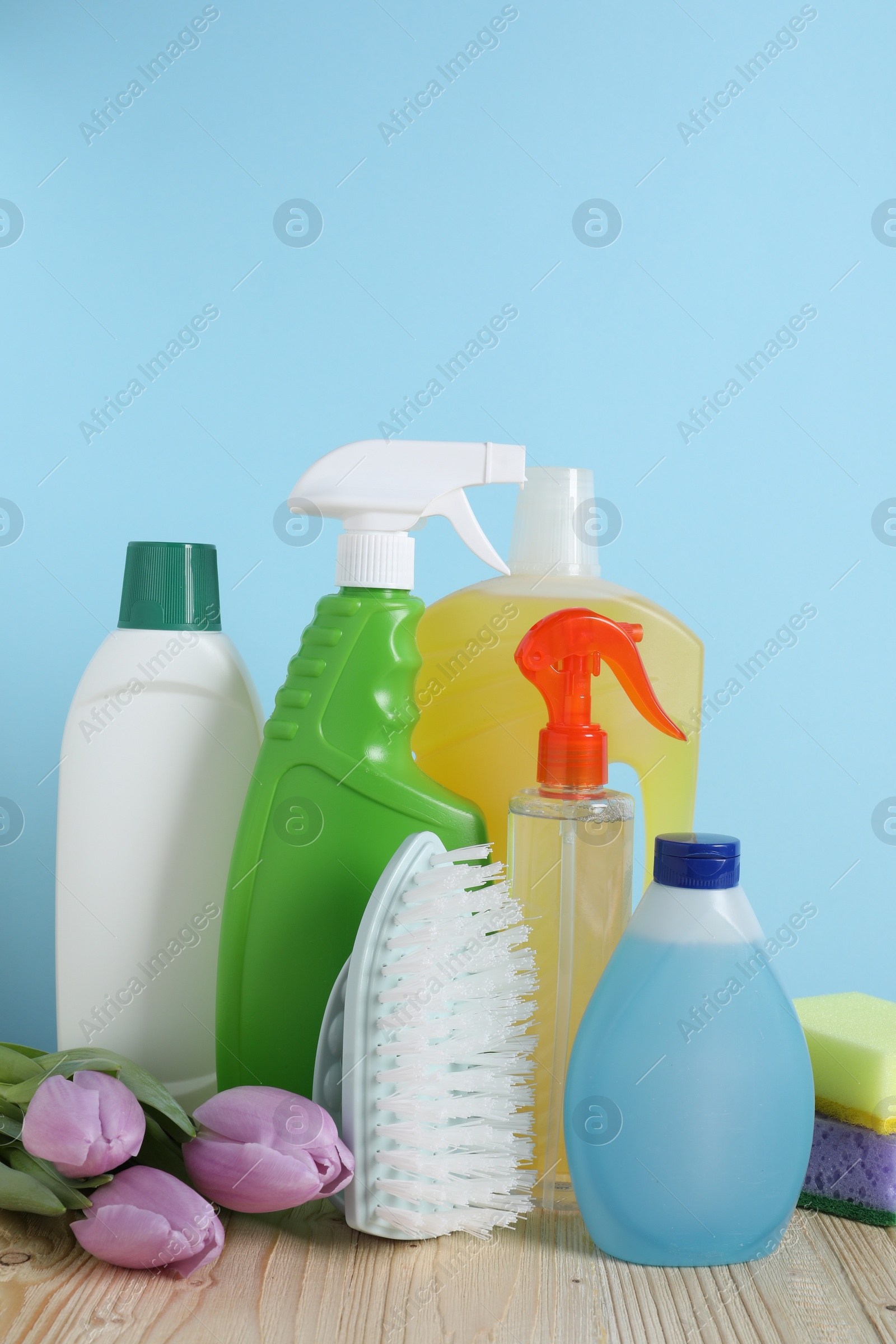 Photo of Spring cleaning. Different detergents, tools and beautiful flowers on wooden table against light blue background