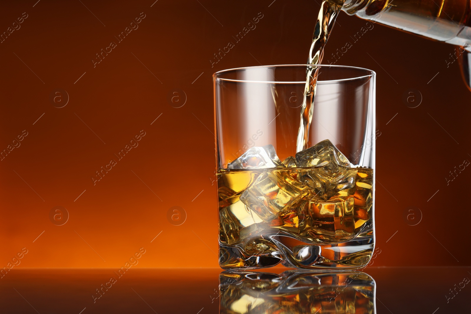 Photo of Pouring whiskey into glass with ice cubes at table against color background, space for text