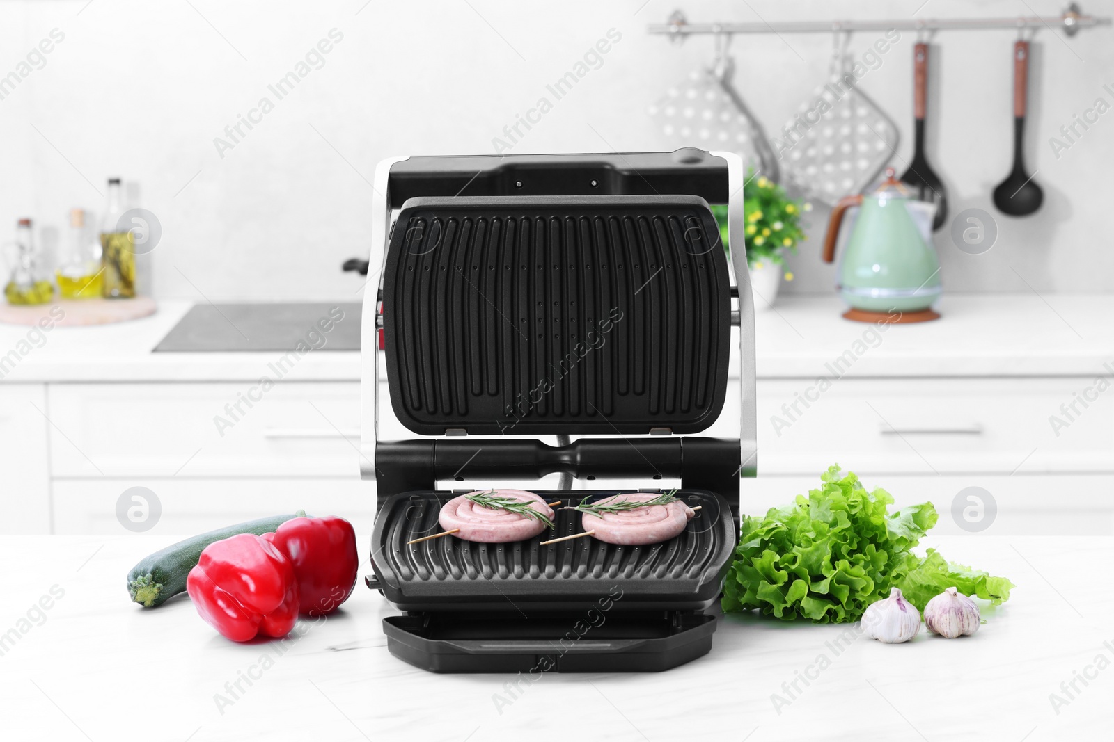 Photo of Electric grill with homemade sausages, rosemary and vegetables on white table in kitchen