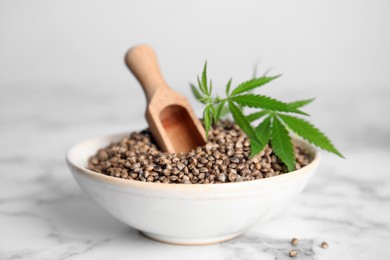 Bowl with hemp seeds, wooden scoop and fresh leaves on white marble table, closeup