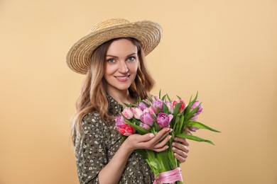 Happy young woman in straw hat holding bouquet of beautiful tulips on beige background