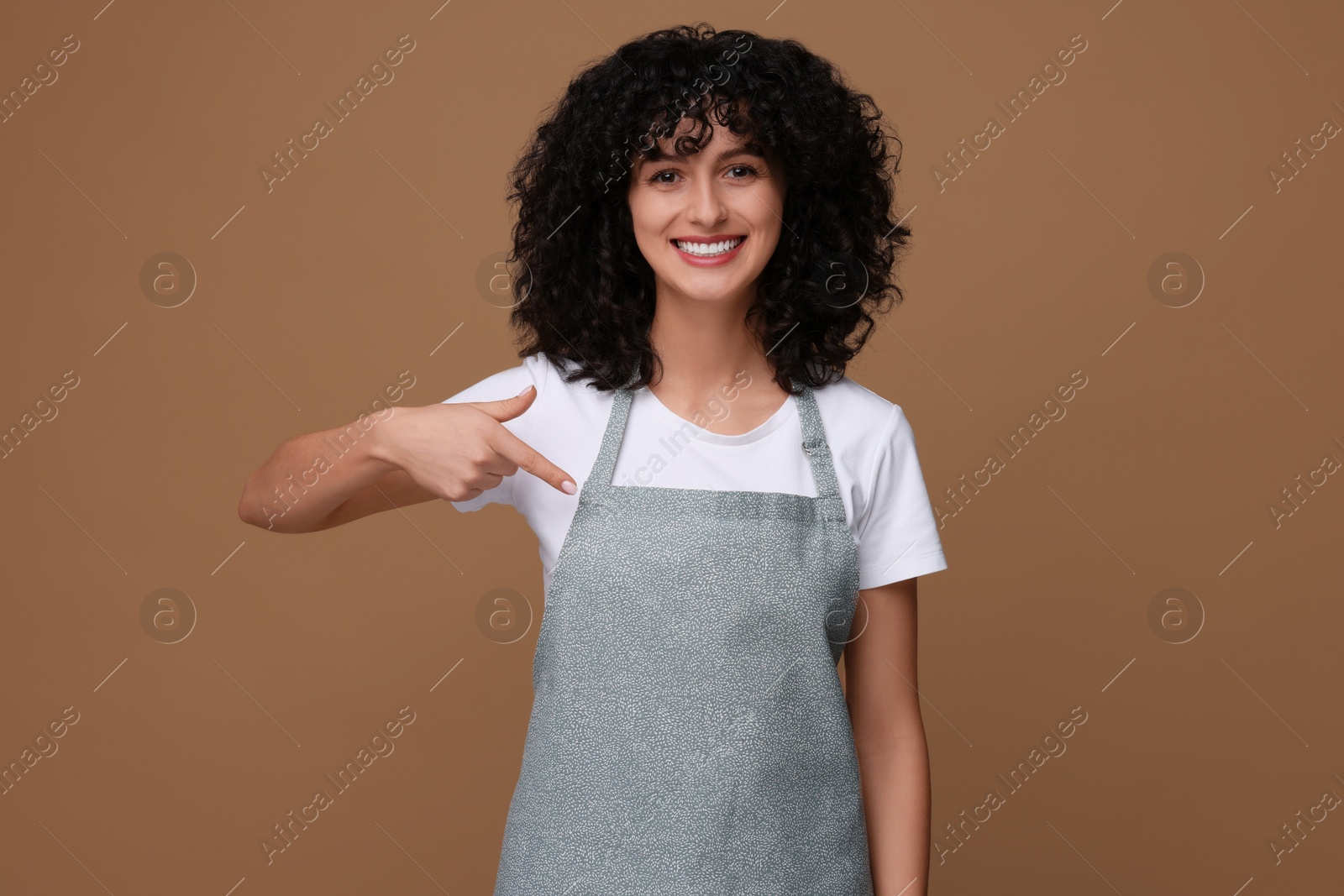 Photo of Happy woman pointing at kitchen apron on brown background. Mockup for design