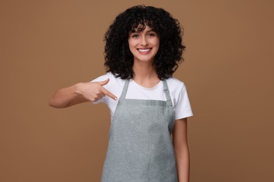 Photo of Happy woman pointing at kitchen apron on brown background. Mockup for design