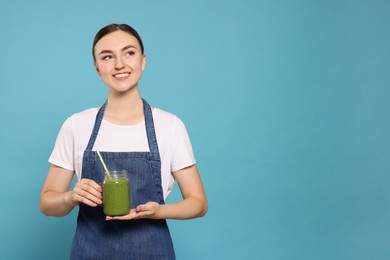 Photo of Beautiful young woman in denim apron with delicious smoothie on light blue background. Space for text