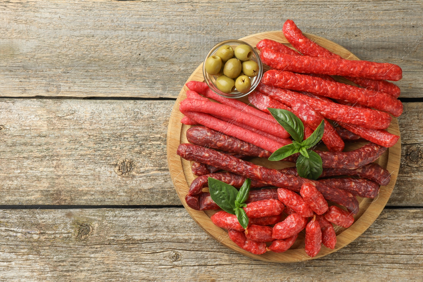 Photo of Different thin dry smoked sausages, basil and olives on wooden table, top view. Space for text