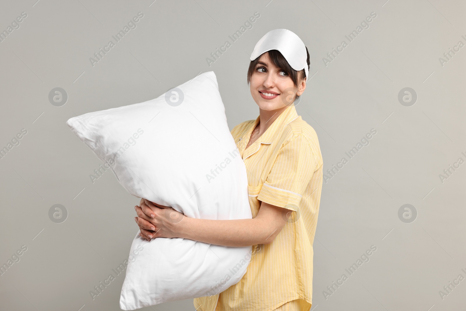 Photo of Happy woman in pyjama and sleep mask holding pillow on grey background