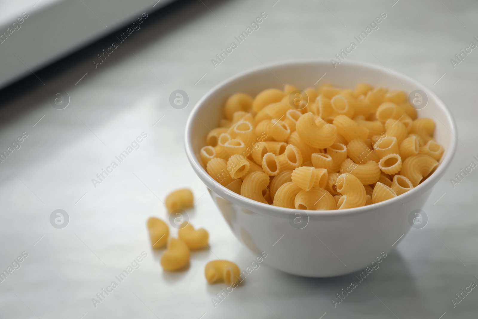 Photo of Bowl with uncooked horns pasta on white marble table, space for text