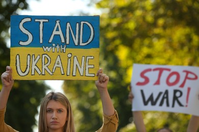 Sad woman holding poster in colors of national flag and words Stand with Ukraine outdoors