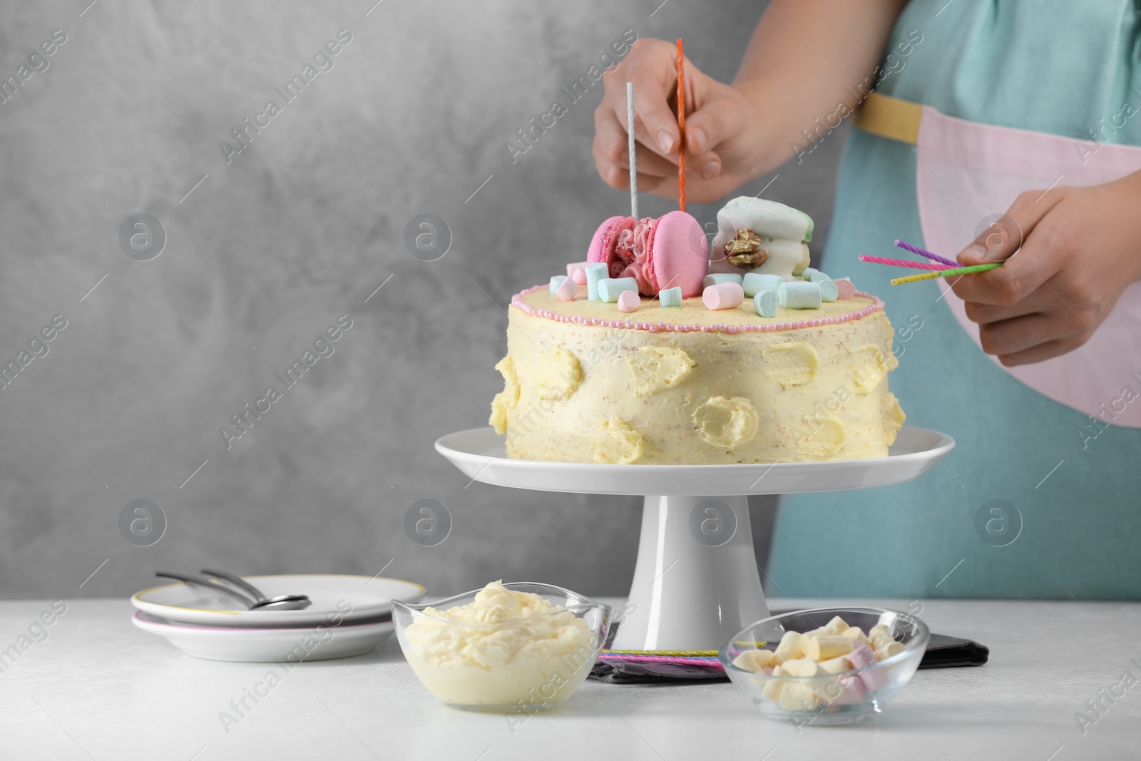 Photo of Woman putting candles on cake decorated with macarons and marshmallows at white table, closeup. Space for text
