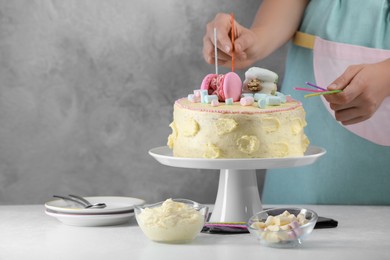 Photo of Woman putting candles on cake decorated with macarons and marshmallows at white table, closeup. Space for text