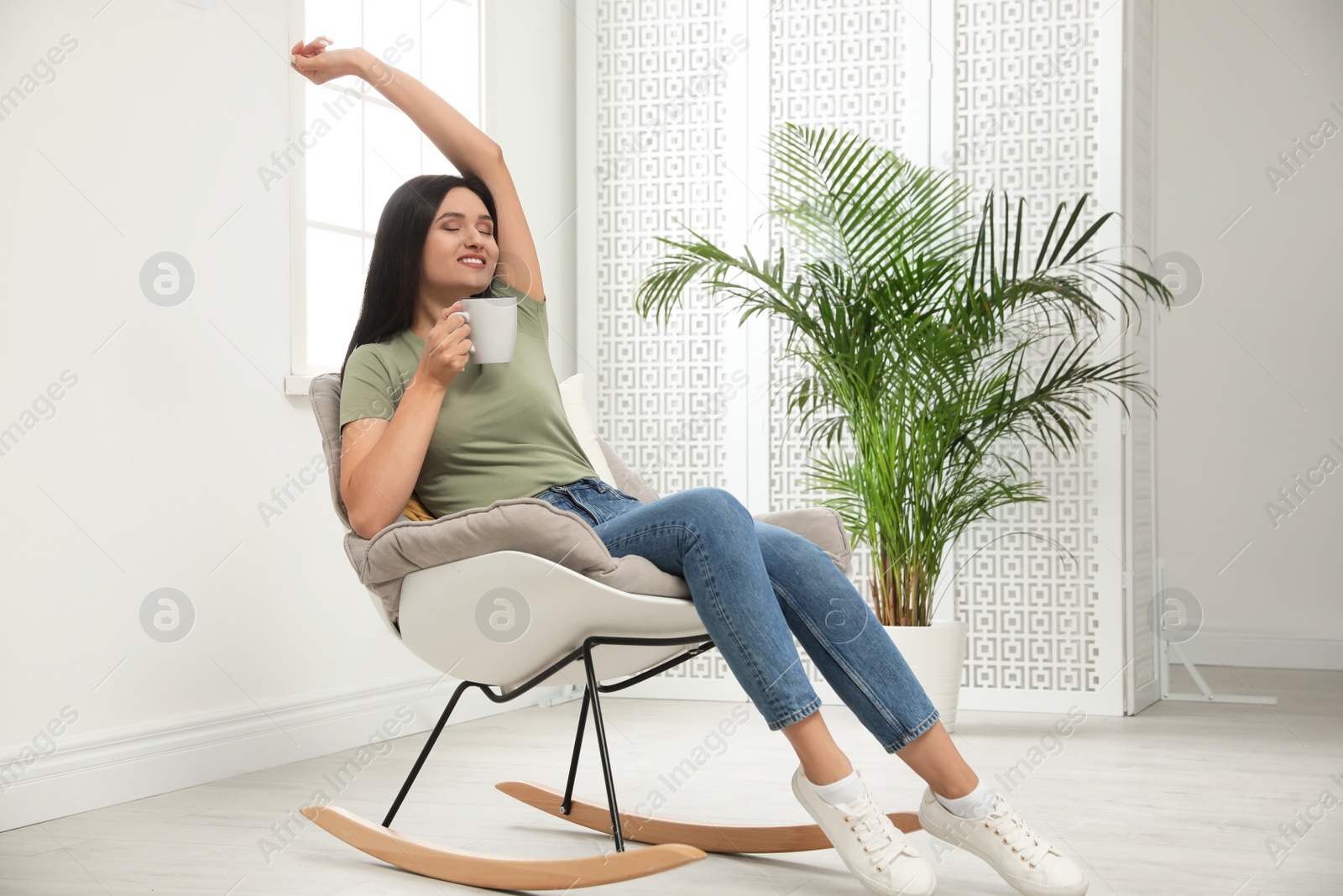 Photo of Young woman with cup of drink relaxing in rocking  chair at home