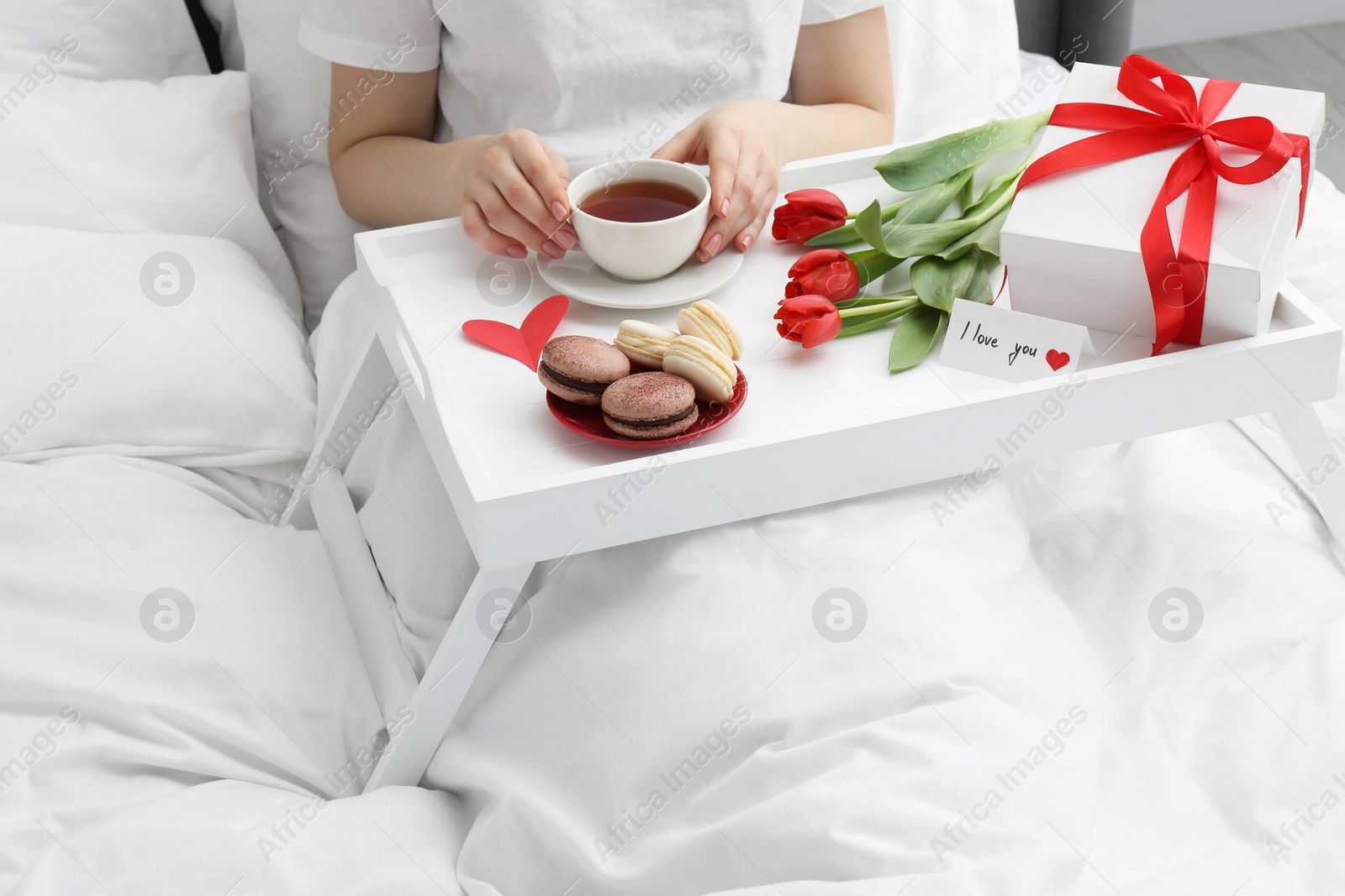 Photo of Tasty breakfast served in bed. Woman with tea, macarons, gift box, flowers and I Love You card at home, closeup