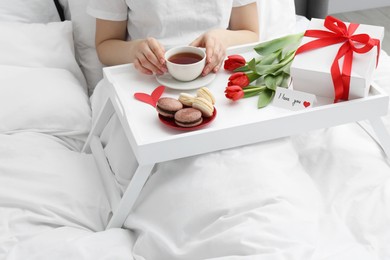 Photo of Tasty breakfast served in bed. Woman with tea, macarons, gift box, flowers and I Love You card at home, closeup