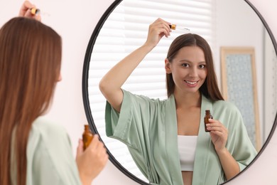 Photo of Beautiful woman applying serum onto hair near mirror indoors