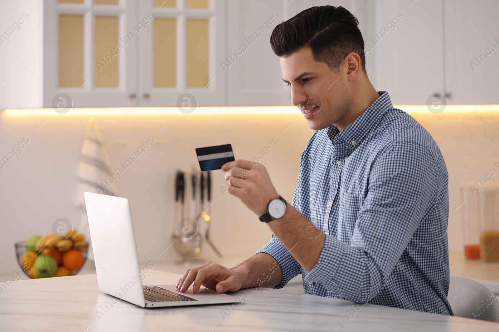 Photo of Man using laptop and credit card for online payment at table in kitchen