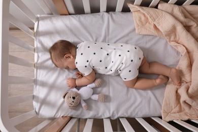 Photo of Adorable little baby with pacifier and toy sleeping in crib indoors, top view