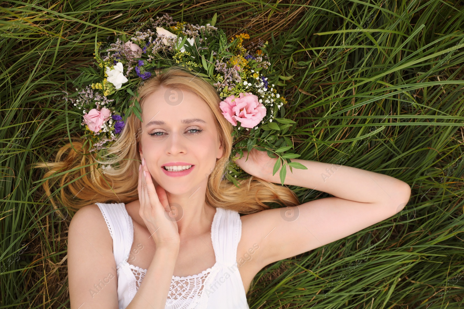 Photo of Young woman wearing wreath made of beautiful flowers on green grass, top view