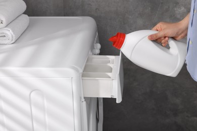 Woman pouring fabric softener from bottle into washing machine near grey wall, closeup
