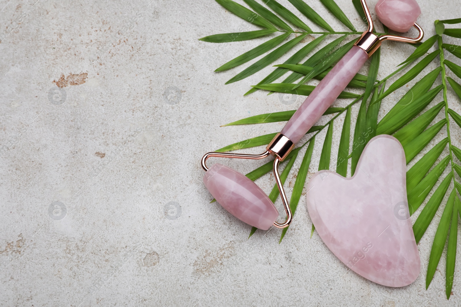 Photo of Gua sha stone, face roller and green leaves on light table, flat lay. Space for text