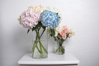 Beautiful hydrangea and rose flowers in vases on white bedside table indoors