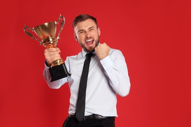 Photo of Portrait of happy young businessman with gold trophy cup on red background