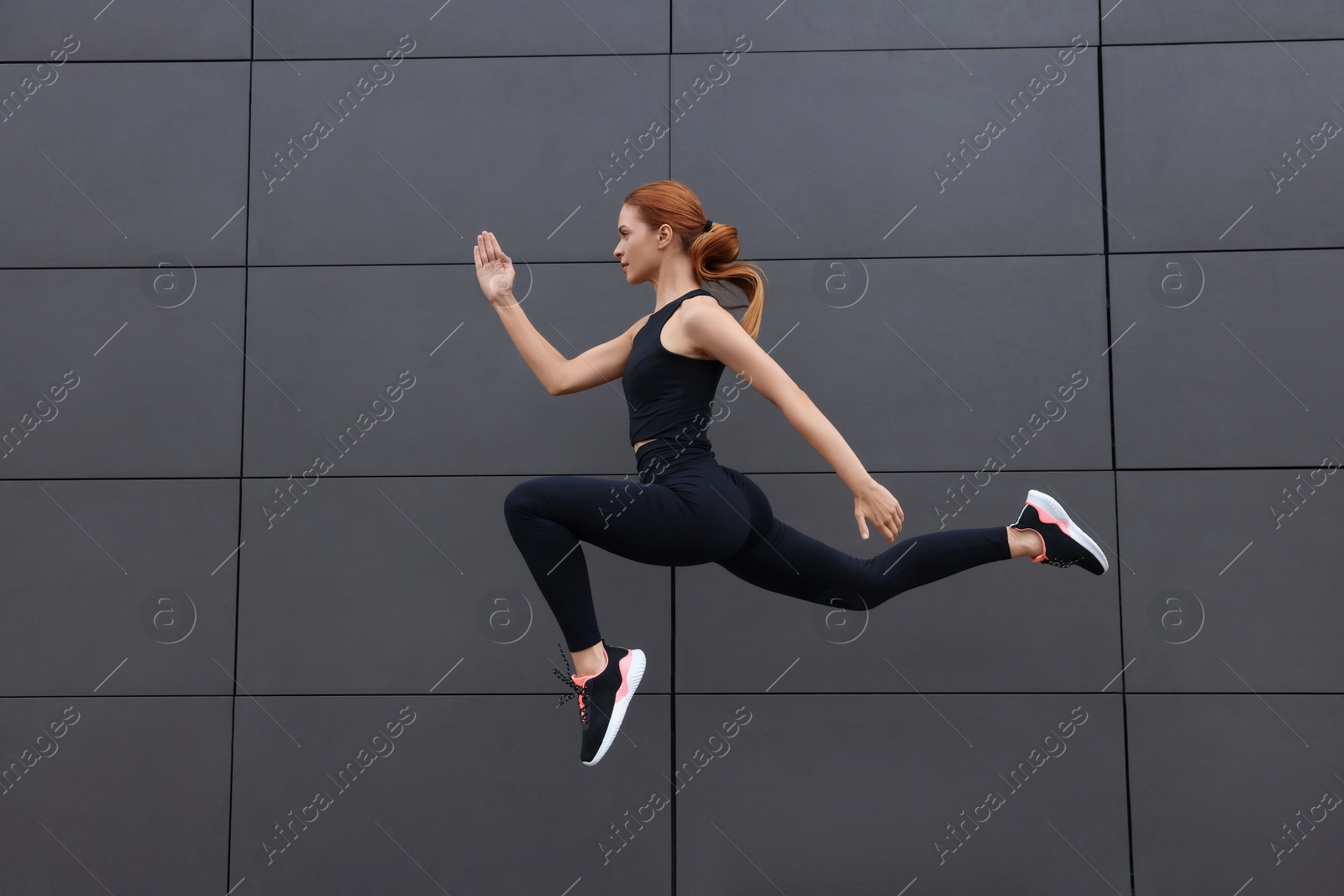 Photo of Beautiful woman in gym clothes jumping near dark grey wall on street