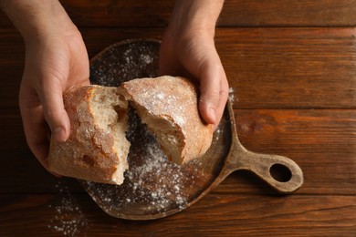 Photo of Man breaking loaf of fresh bread at wooden table, top view