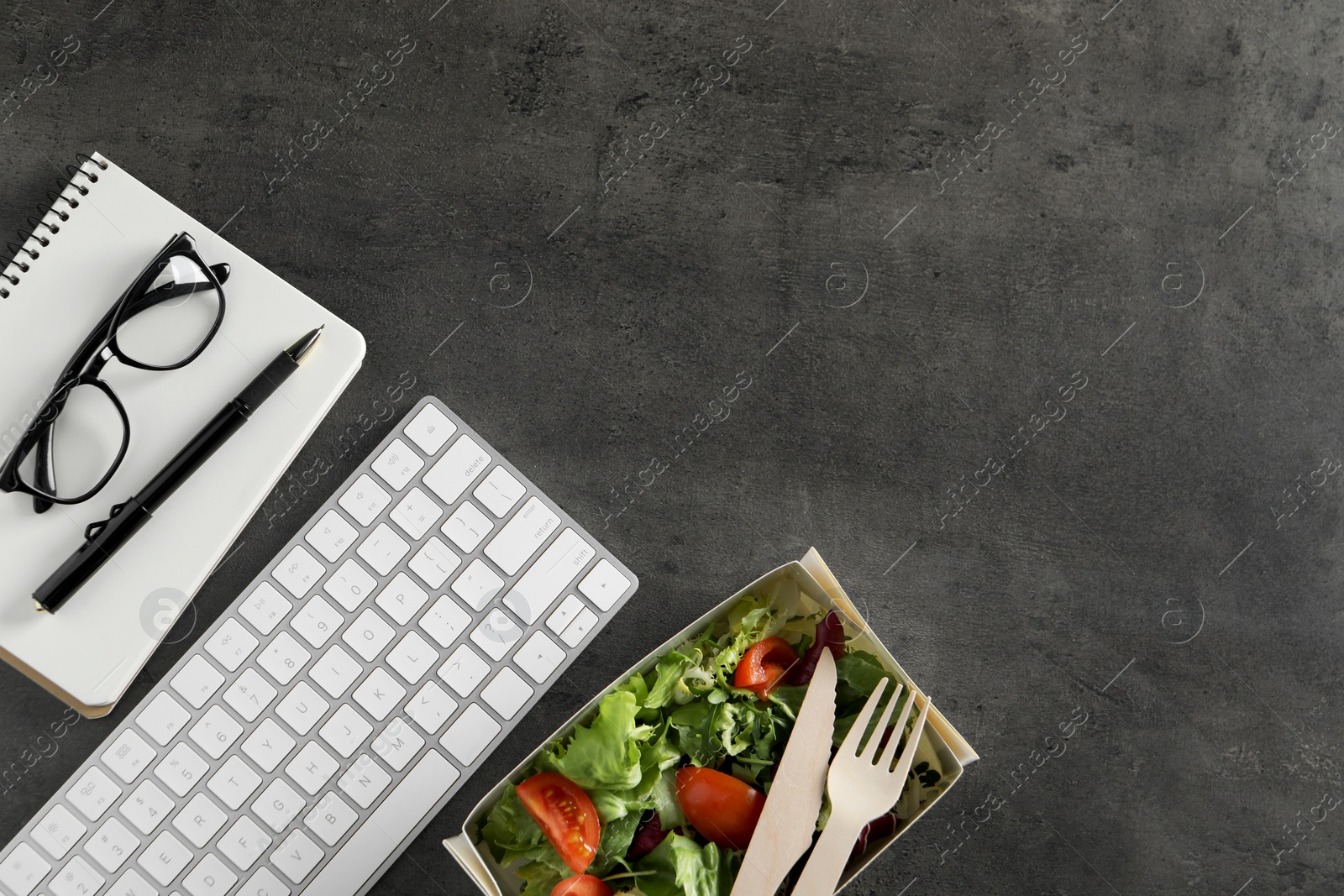 Photo of Container of tasty food, keyboard, glasses, cutlery and notebook on grey table, flat lay with space for text. Business lunch