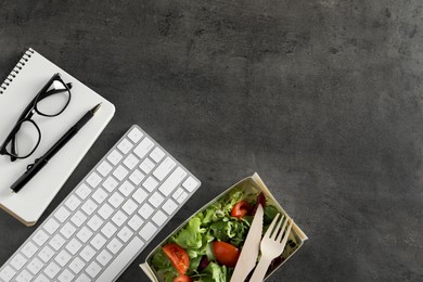 Photo of Container of tasty food, keyboard, glasses, cutlery and notebook on grey table, flat lay with space for text. Business lunch