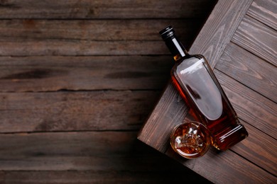 Whiskey with ice cubes in glass and bottle on wooden crate, top view. Space for text