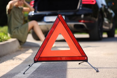 Woman sitting near broken car on roadside outdoors, focus on warning triangle