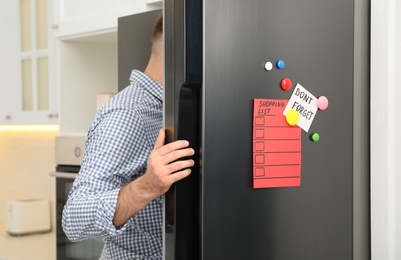 Photo of Man opening refrigerator door with shopping list, note and magnets in kitchen