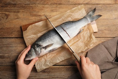 Photo of Woman cutting raw sea bass fish at wooden table, top view