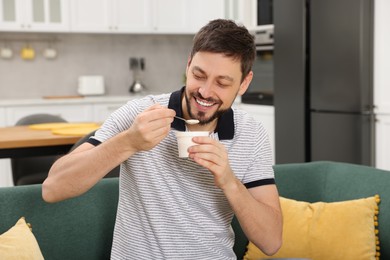 Photo of Handsome man eating tasty yogurt on sofa in kitchen