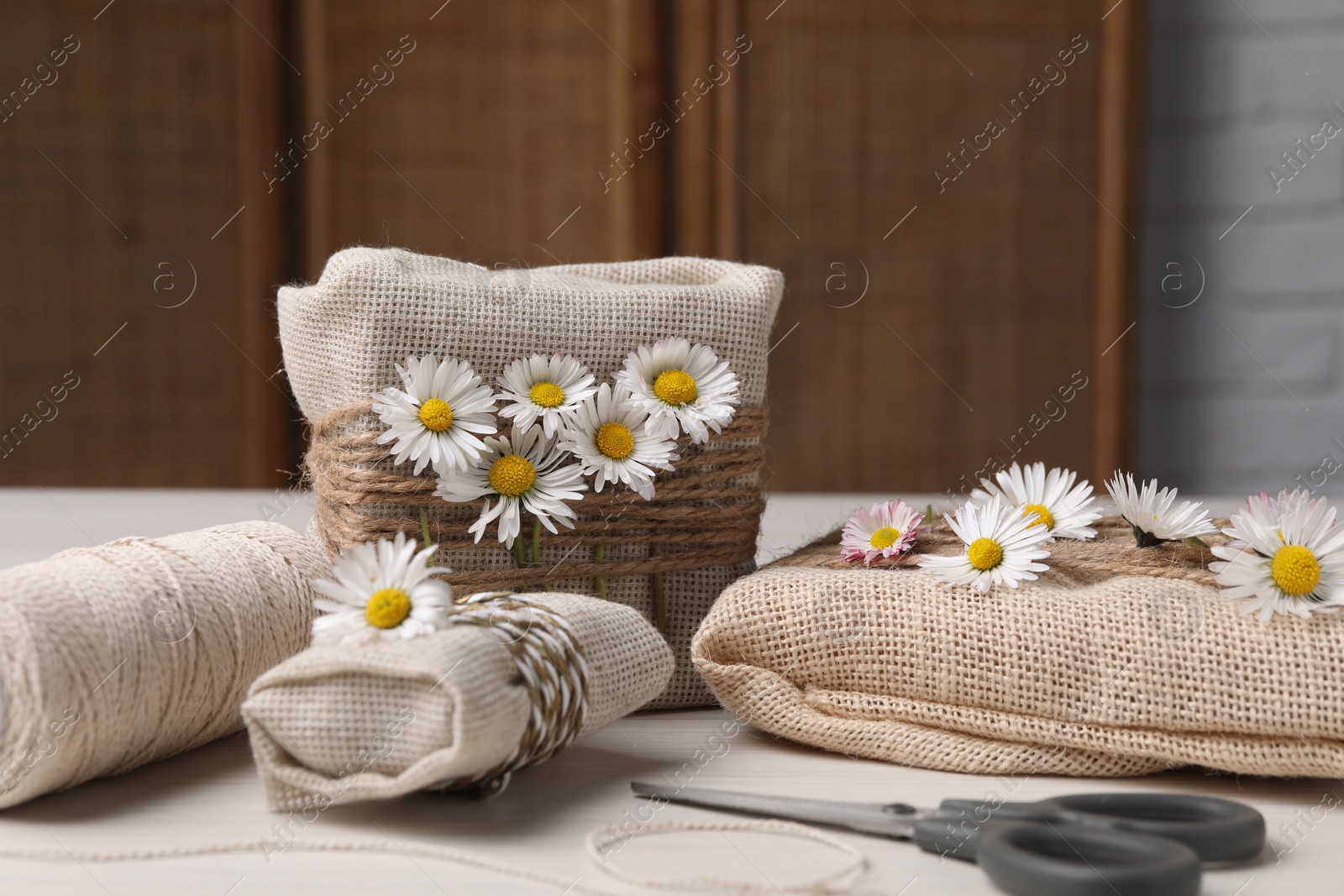 Photo of Gifts packed in burlap fabric, beautiful chamomiles, ropes, spool of threads and scissors on white table