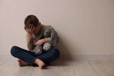 Child abuse. Upset boy with toy bunny sitting on floor near beige wall indoors, space for text