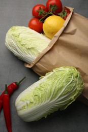 Photo of Paper bag with fresh Chinese cabbages, lemon, tomatoes and chili pepper on grey textured table, flat lay