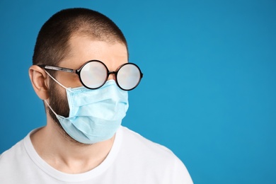Photo of Man with foggy glasses caused by wearing disposable mask on blue background, space for text. Protective measure during coronavirus pandemic