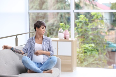 Trendy pregnant woman with tattoo on sofa indoors