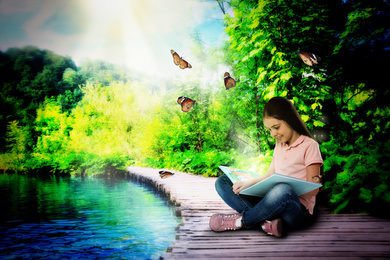 Cute little girl reading magic book on wooden bridge near lake 