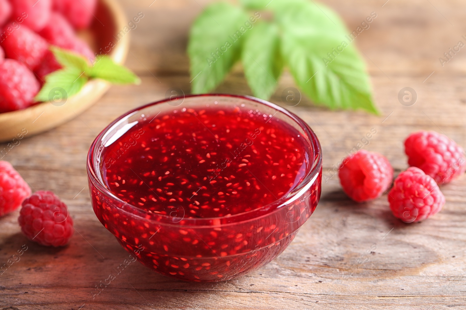 Photo of Bowl of sweet jam with ripe raspberries on wooden table, closeup