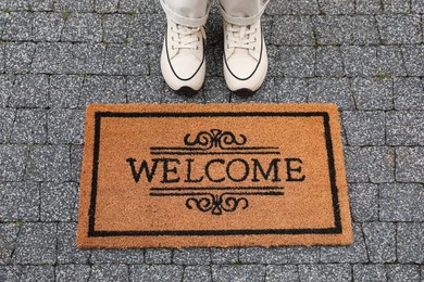 Photo of Woman near doormat with word Welcome on pavement outdoors, closeup