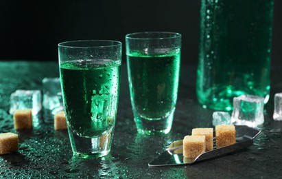 Photo of Absinthe in shot glasses, spoon, brown sugar and ice cubes on gray table against dark background, closeup. Alcoholic drink