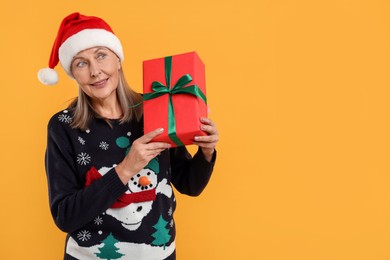 Photo of Senior woman in Christmas sweater and Santa hat holding gift on orange background. Space for text