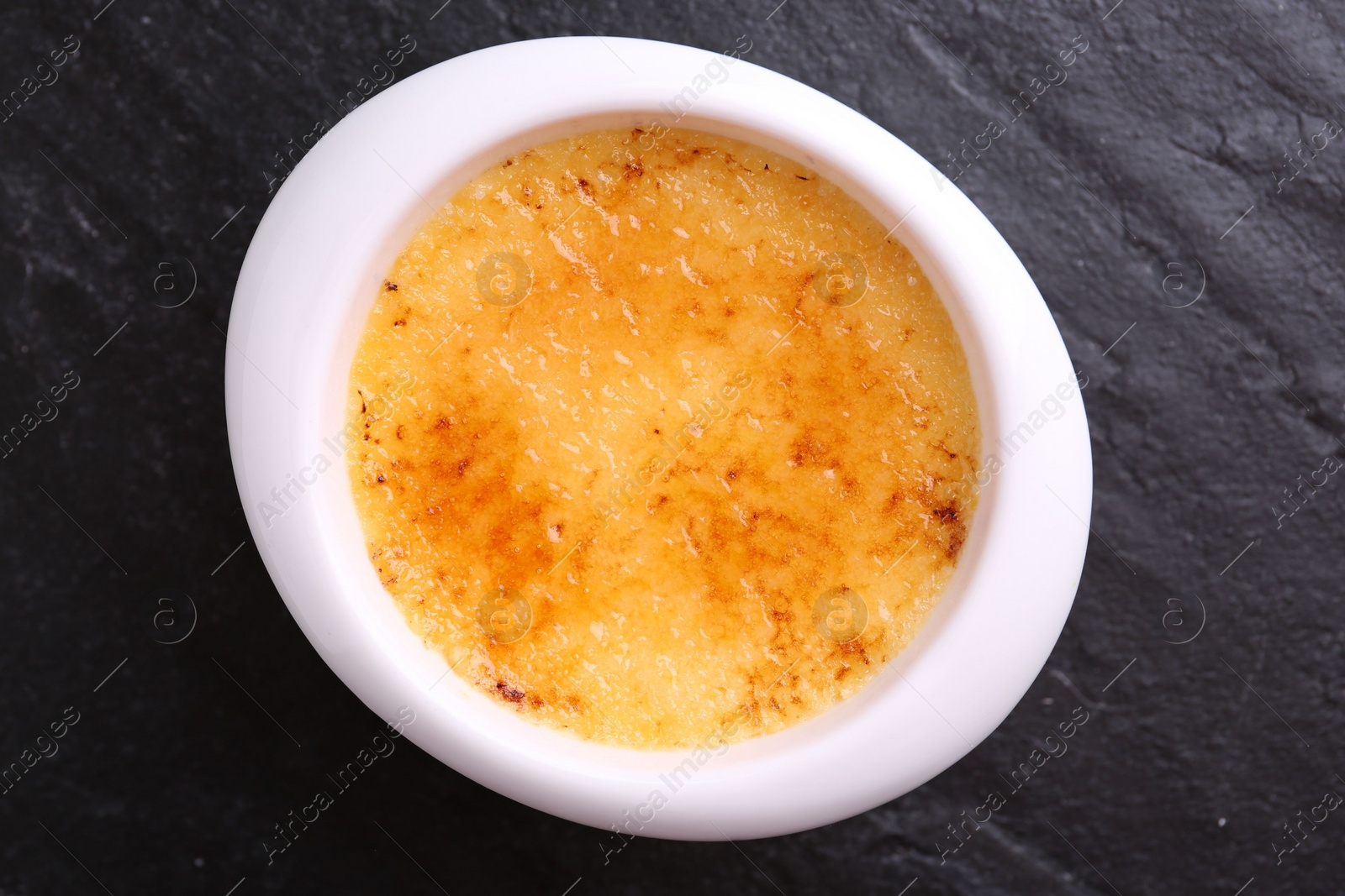 Photo of Delicious creme brulee in bowl on dark gray textured table, top view