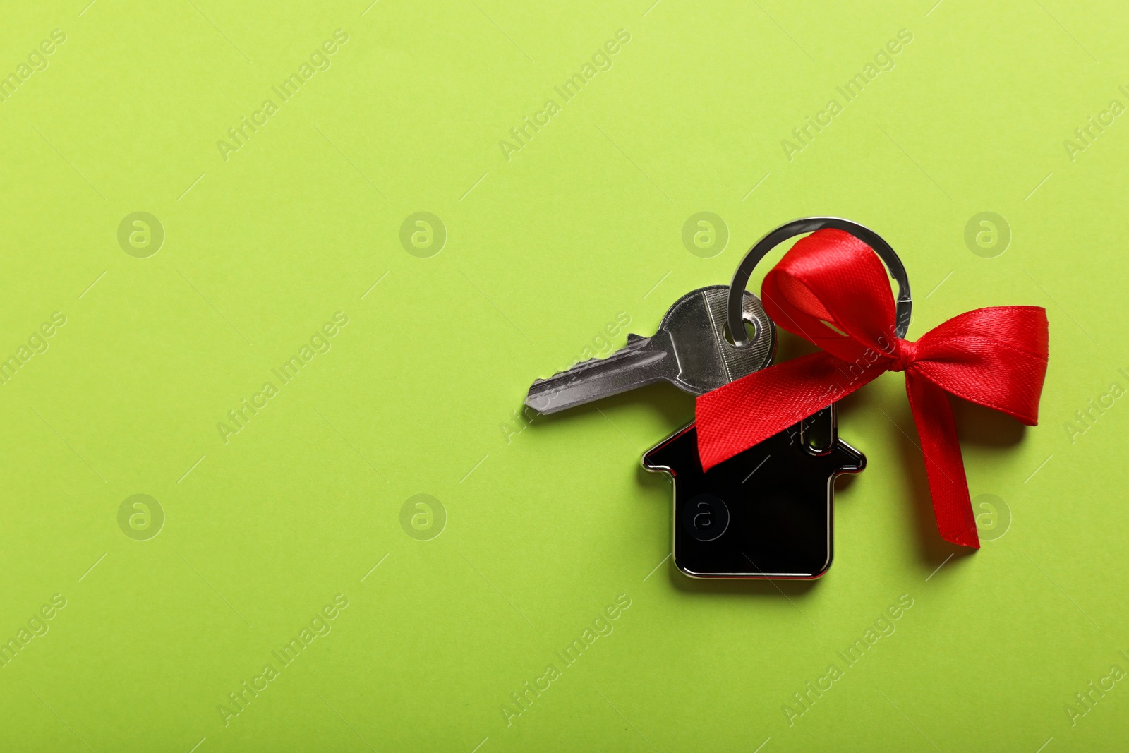 Photo of Key with trinket in shape of house and red bow on light green background, top view. Space for text. Housewarming party