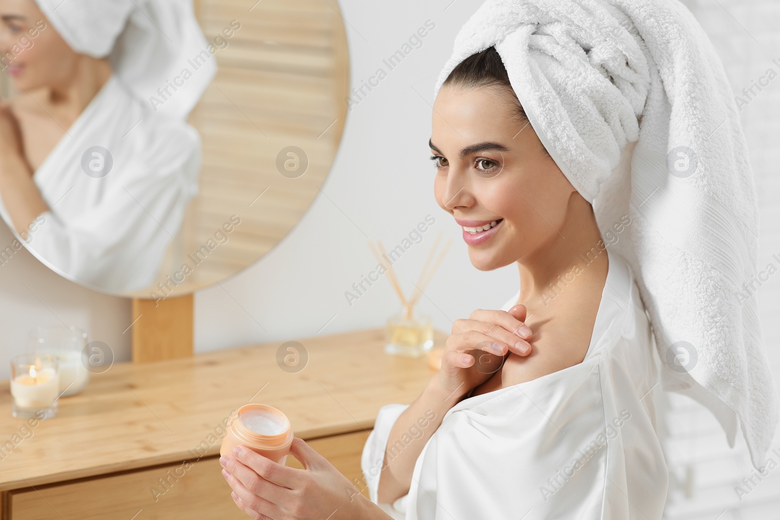 Photo of Beautiful happy woman in stylish bathrobe applying cream on shoulder in bathroom