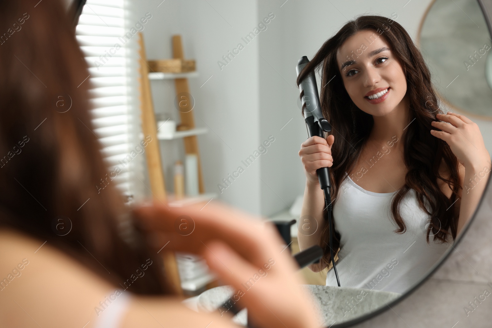 Photo of Smiling woman using curling hair iron near mirror in bathroom