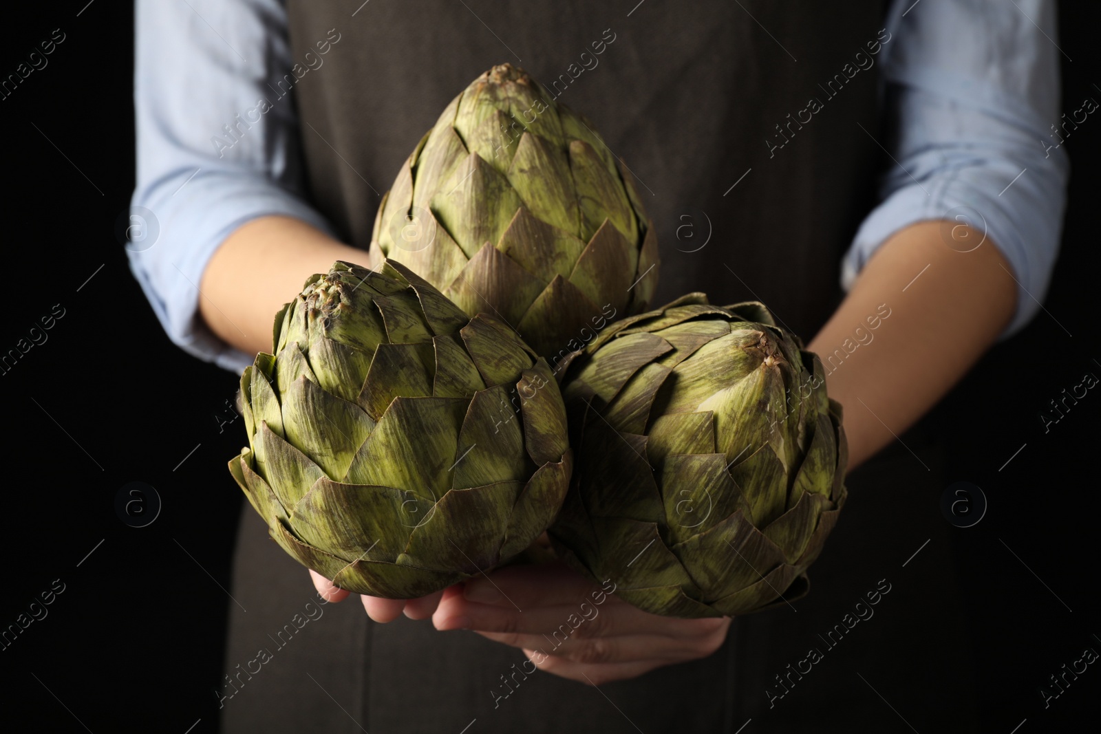 Photo of Woman holding fresh raw artichokes on black background, closeup