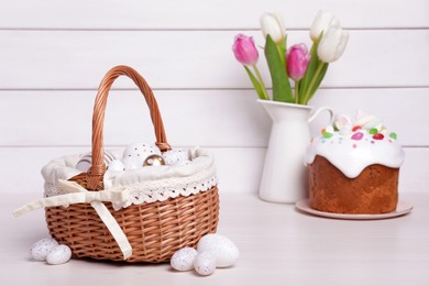 Wicker basket with festively decorated eggs, beautiful tulips and traditional Easter cake on white wooden table